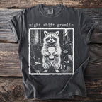 Dark gray t-shirt featuring "night shift gremlin" text above a raccoon holding a coffee cup.