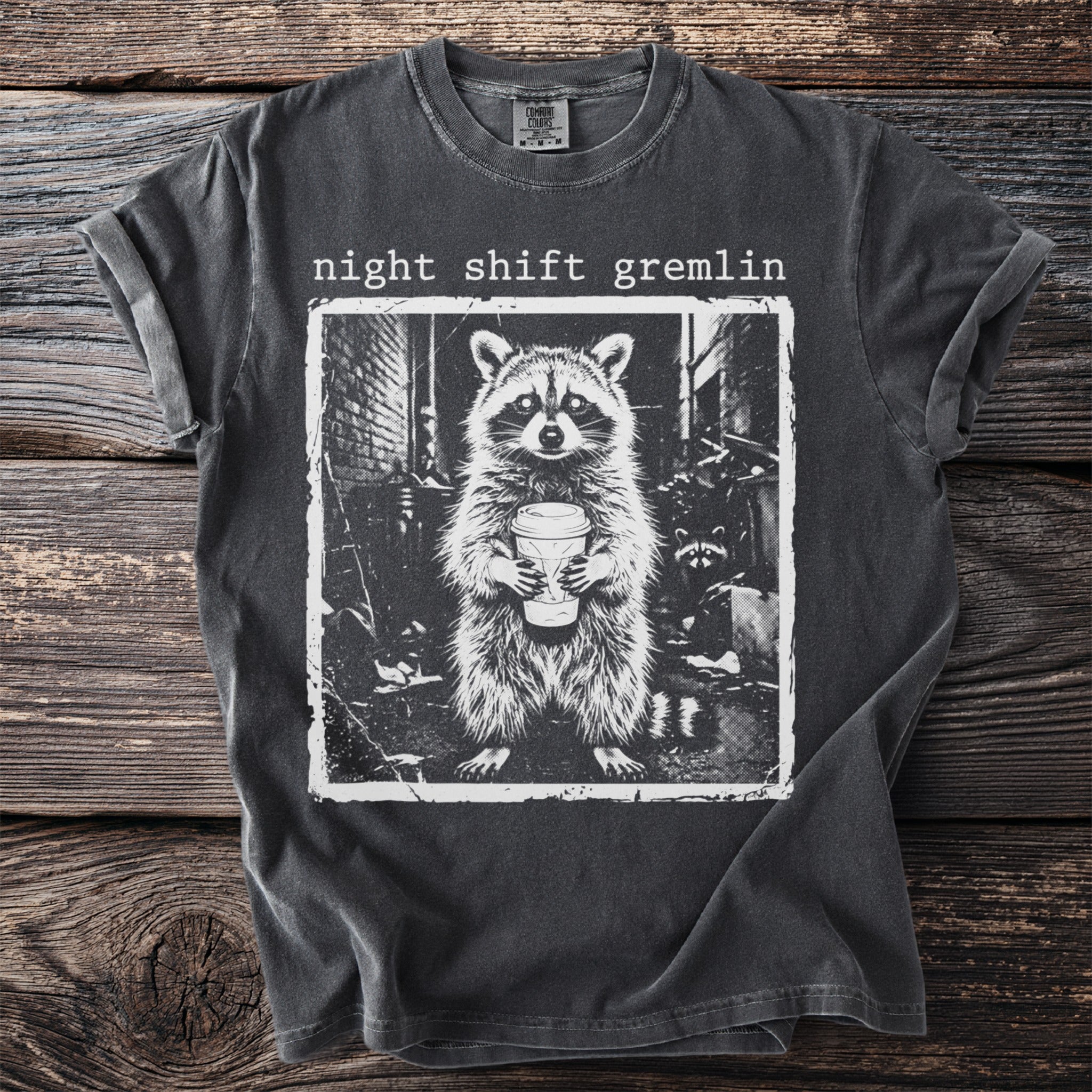 Dark gray t-shirt featuring "night shift gremlin" text above a raccoon holding a coffee cup.