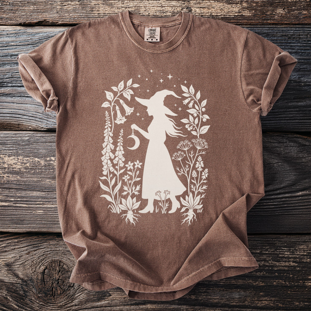 Brown t-shirt with silhouette of witch holding moon and surrounded by botanical illustrations.