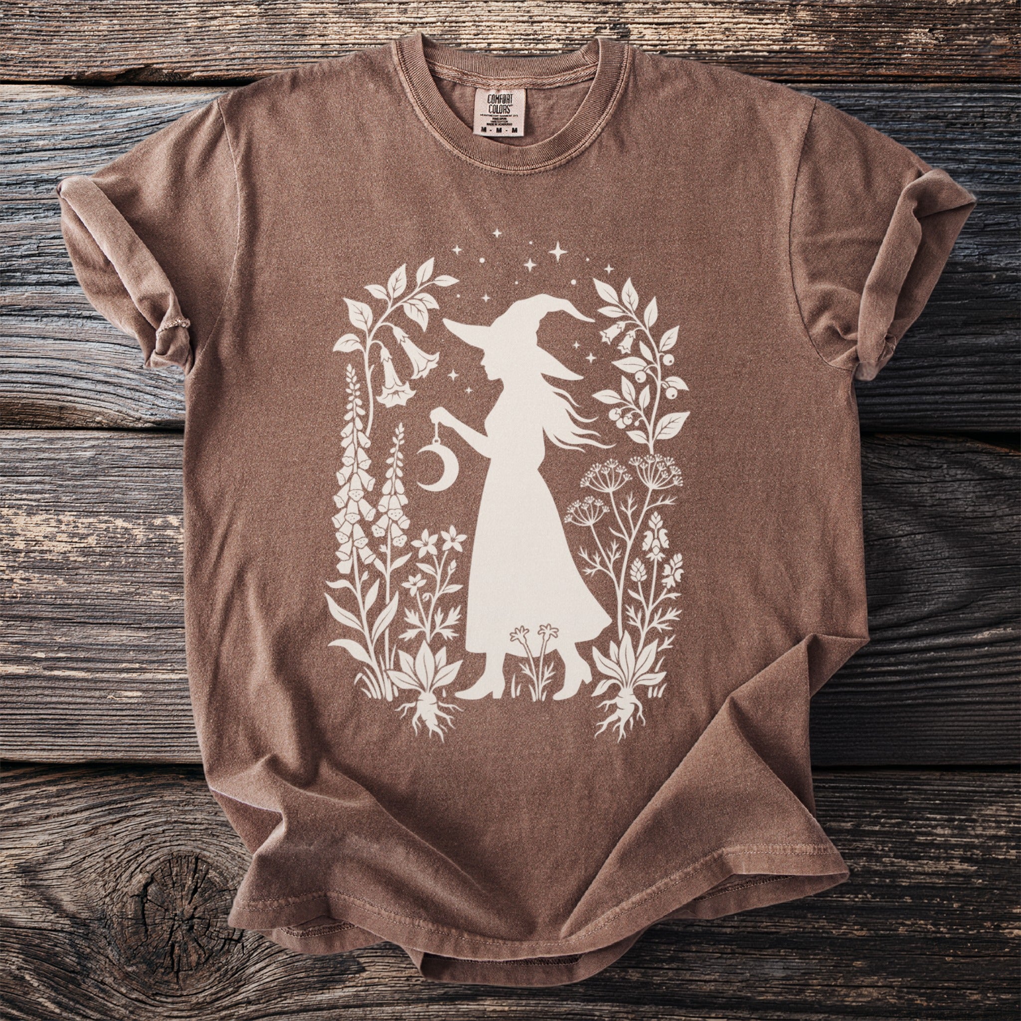 Brown t-shirt with silhouette of witch holding moon and surrounded by botanical illustrations.