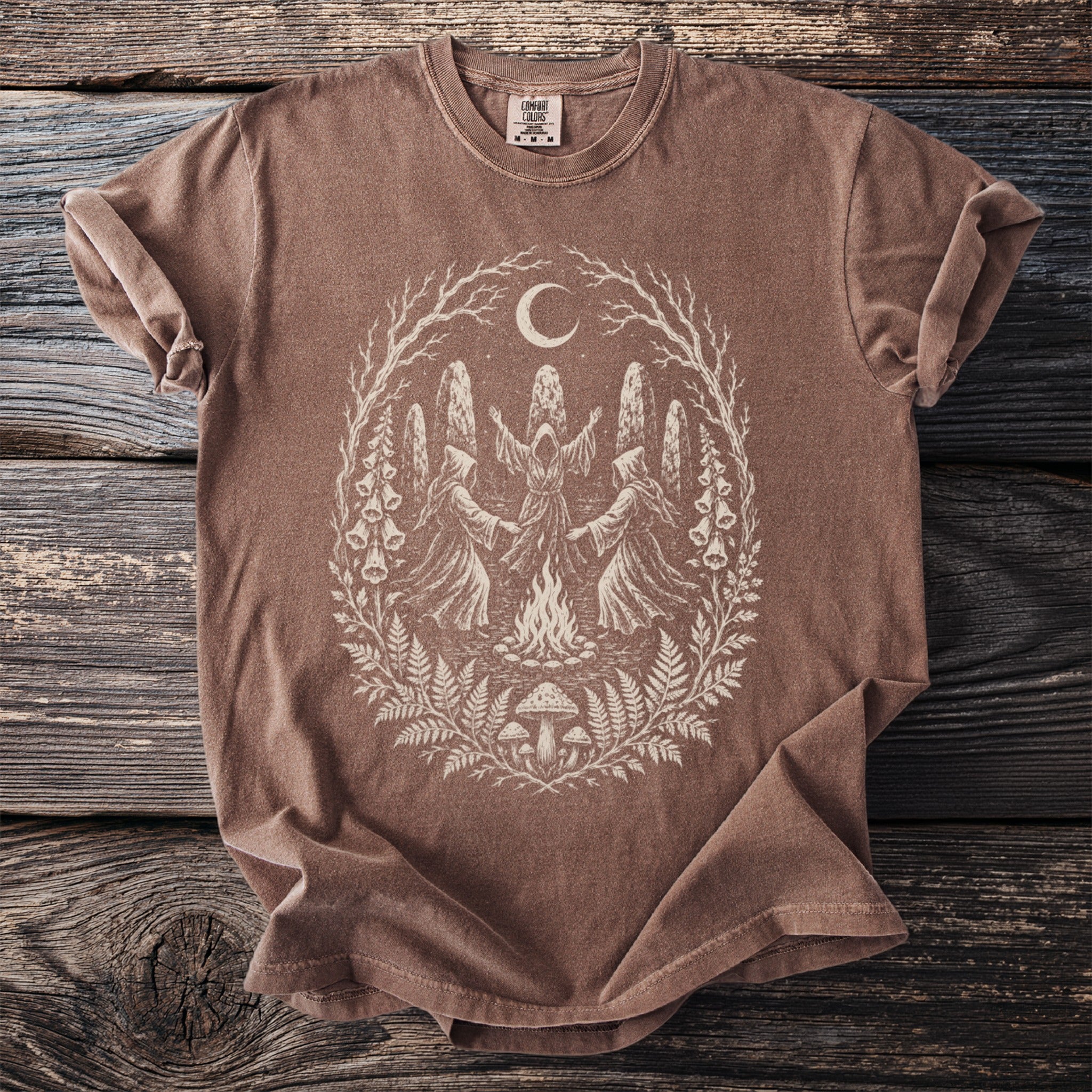 Brown t-shirt with a graphic of robed figures dancing around a campfire under a crescent moon.