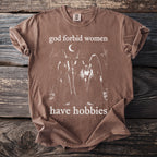 Brown t-shirt with a graphic of three witches, a moon, and text "god forbid women have hobbies".