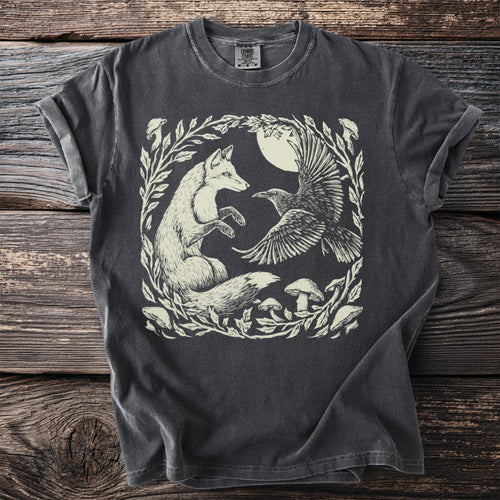 T-shirt with cream illustration of a fox and raven in moonlit forest with mushrooms.