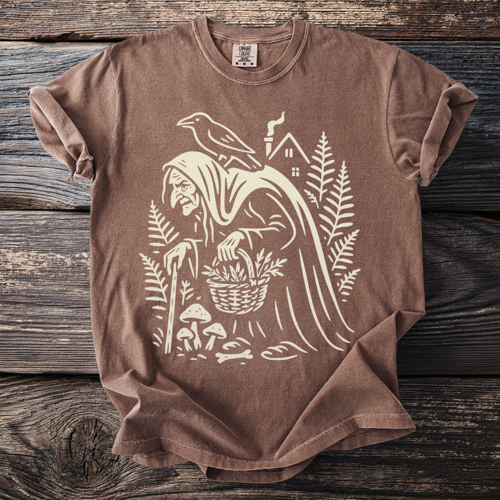 Brown t-shirt with a graphic of a witch, raven, house, mushrooms, and basket.