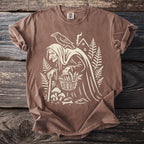 Brown t-shirt with a graphic of a witch, raven, house, mushrooms, and basket.