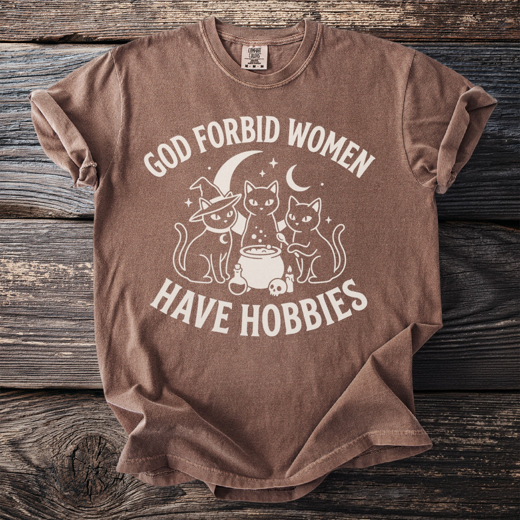 Brown t-shirt with white graphic of three cats, cauldron, and moon, reading "God forbid women have hobbies".