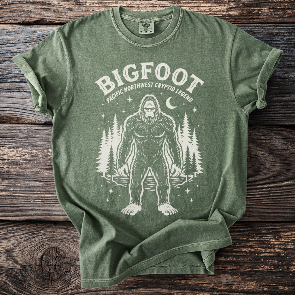 Green t-shirt with Bigfoot and trees graphic and text "BIGFOOT PACIFIC NORTHWEST CRYPTID LEGEND".