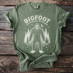 Green t-shirt with Bigfoot and trees graphic and text "BIGFOOT PACIFIC NORTHWEST CRYPTID LEGEND".