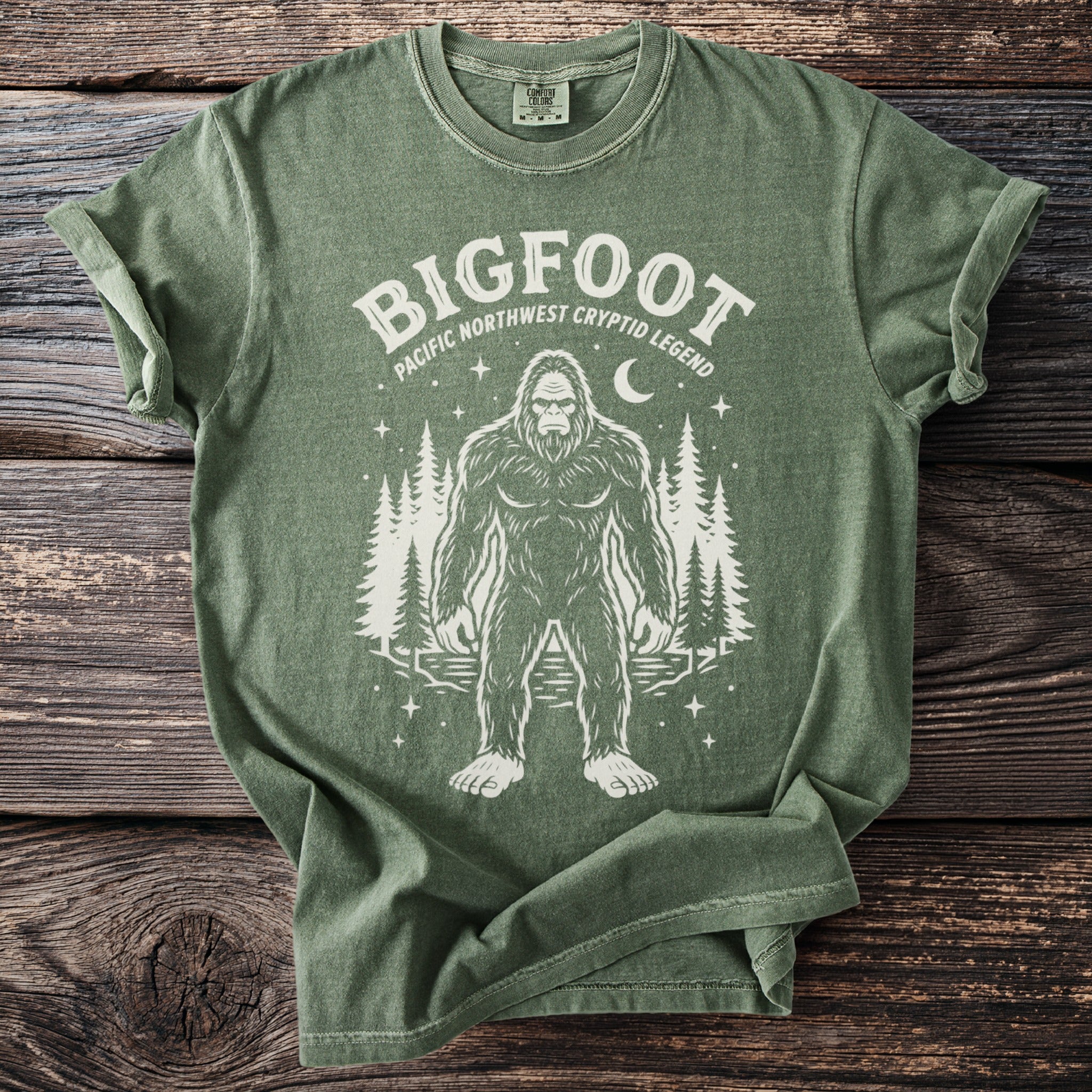 Green t-shirt with Bigfoot and trees graphic and text "BIGFOOT PACIFIC NORTHWEST CRYPTID LEGEND".