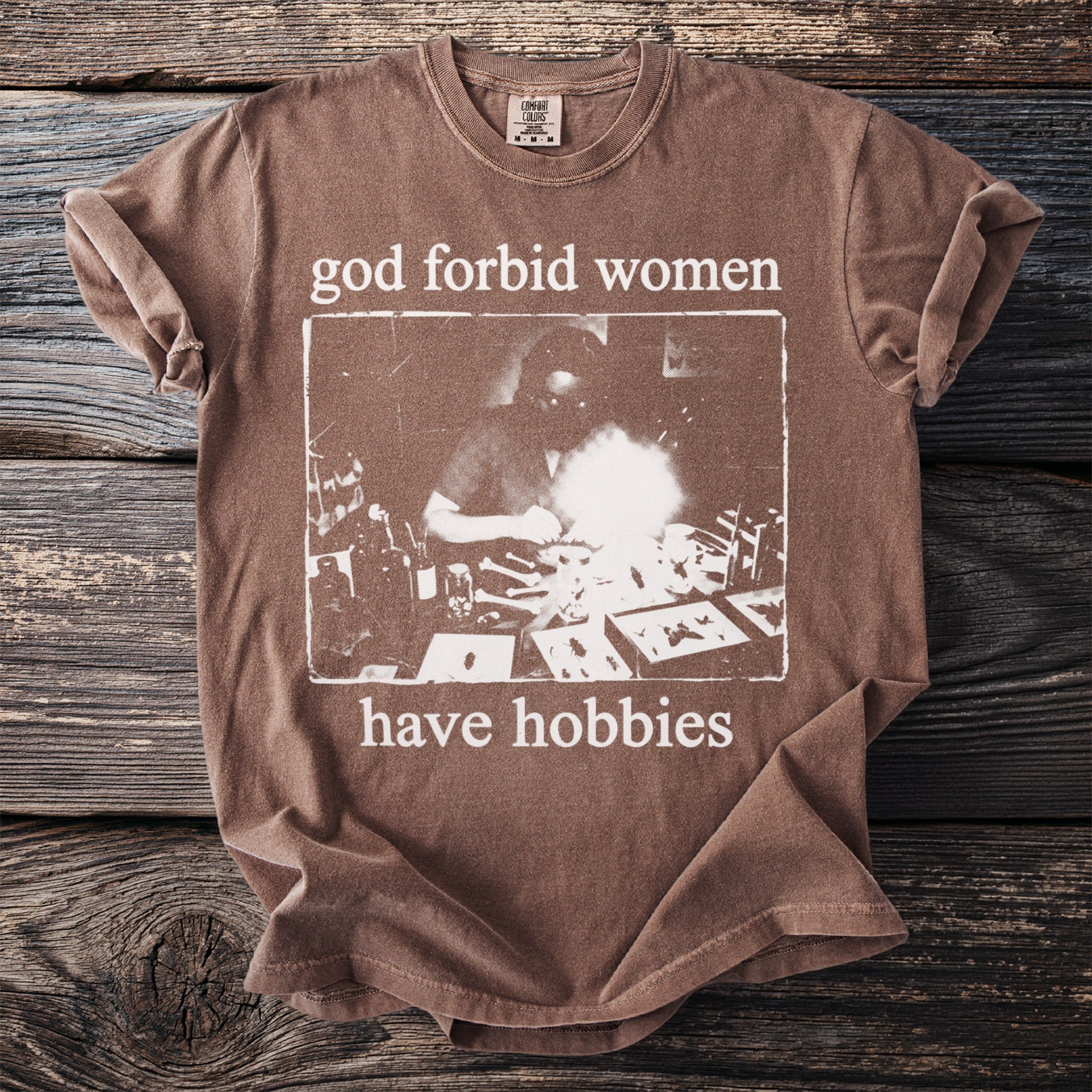 Brown t-shirt with "god forbid women have hobbies" text and vintage illustration.