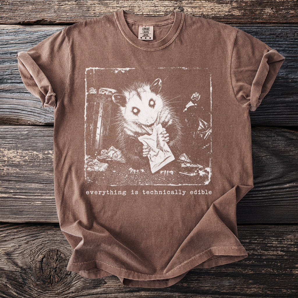 Brown t-shirt with graphic of an opossum eating from a wrapper and text "everything is technically edible".