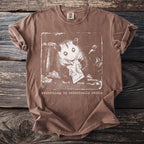 Brown t-shirt with graphic of an opossum eating from a wrapper and text "everything is technically edible".