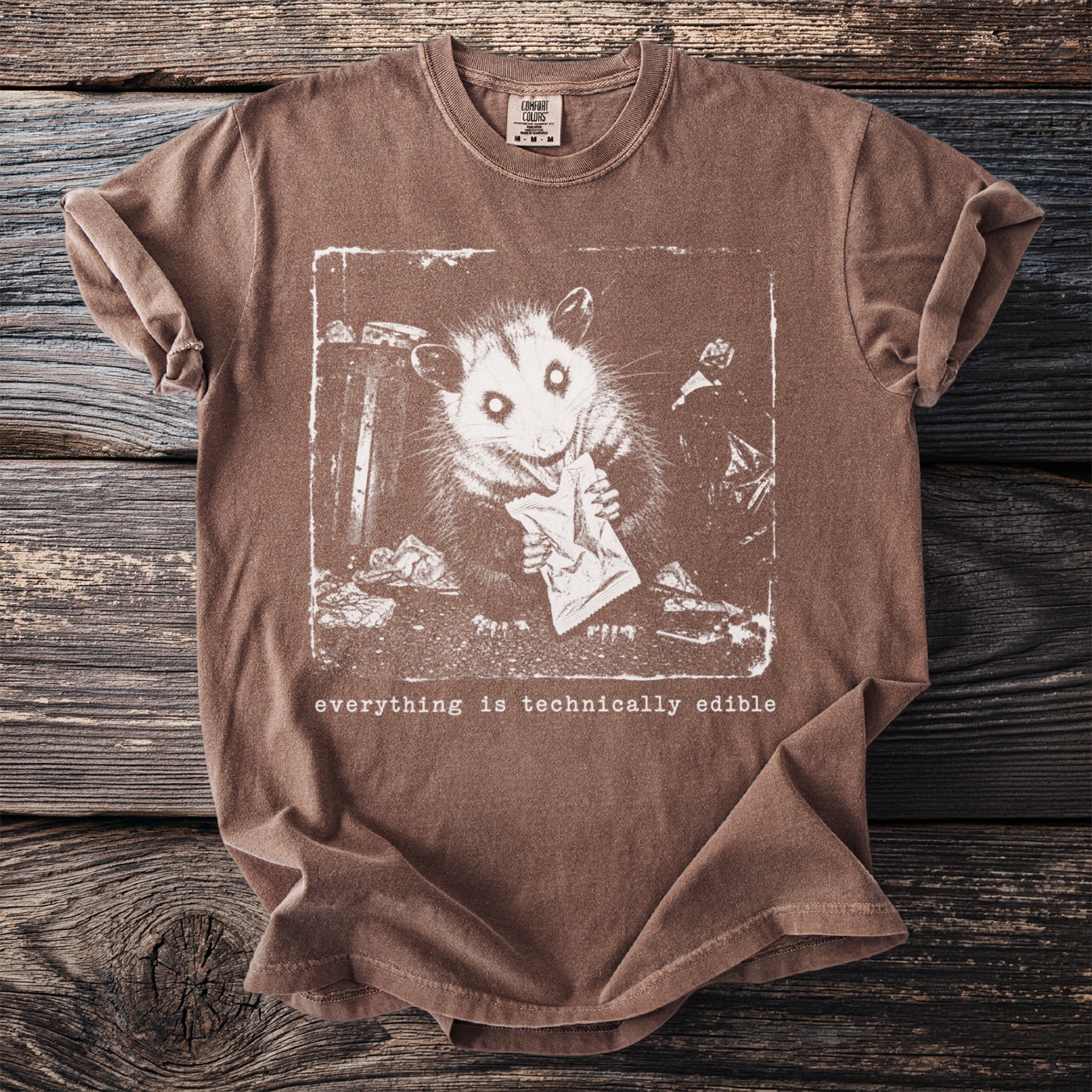 Brown t-shirt with graphic of an opossum eating from a wrapper and text "everything is technically edible".