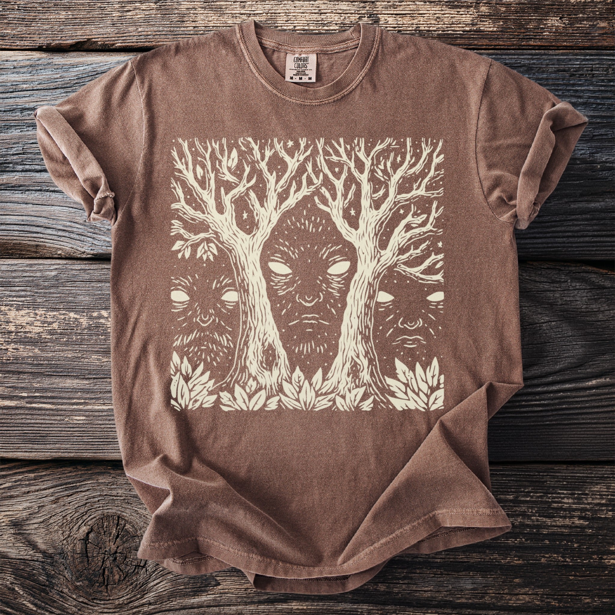 Brown t-shirt with a cream linocut print of a face between two trees.
