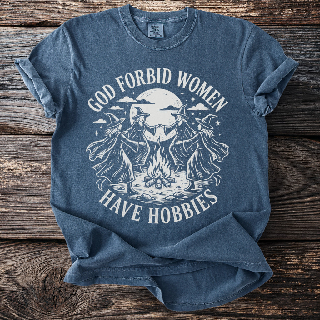 Blue t-shirt with vintage graphic of witches dancing around a bonfire, text reads "God forbid women have hobbies".