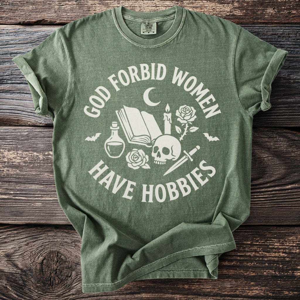 Green t-shirt with white text "God Forbid Women Have Hobbies" and gothic graphic elements.