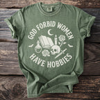 Green t-shirt with white text "God Forbid Women Have Hobbies" and gothic graphic elements.