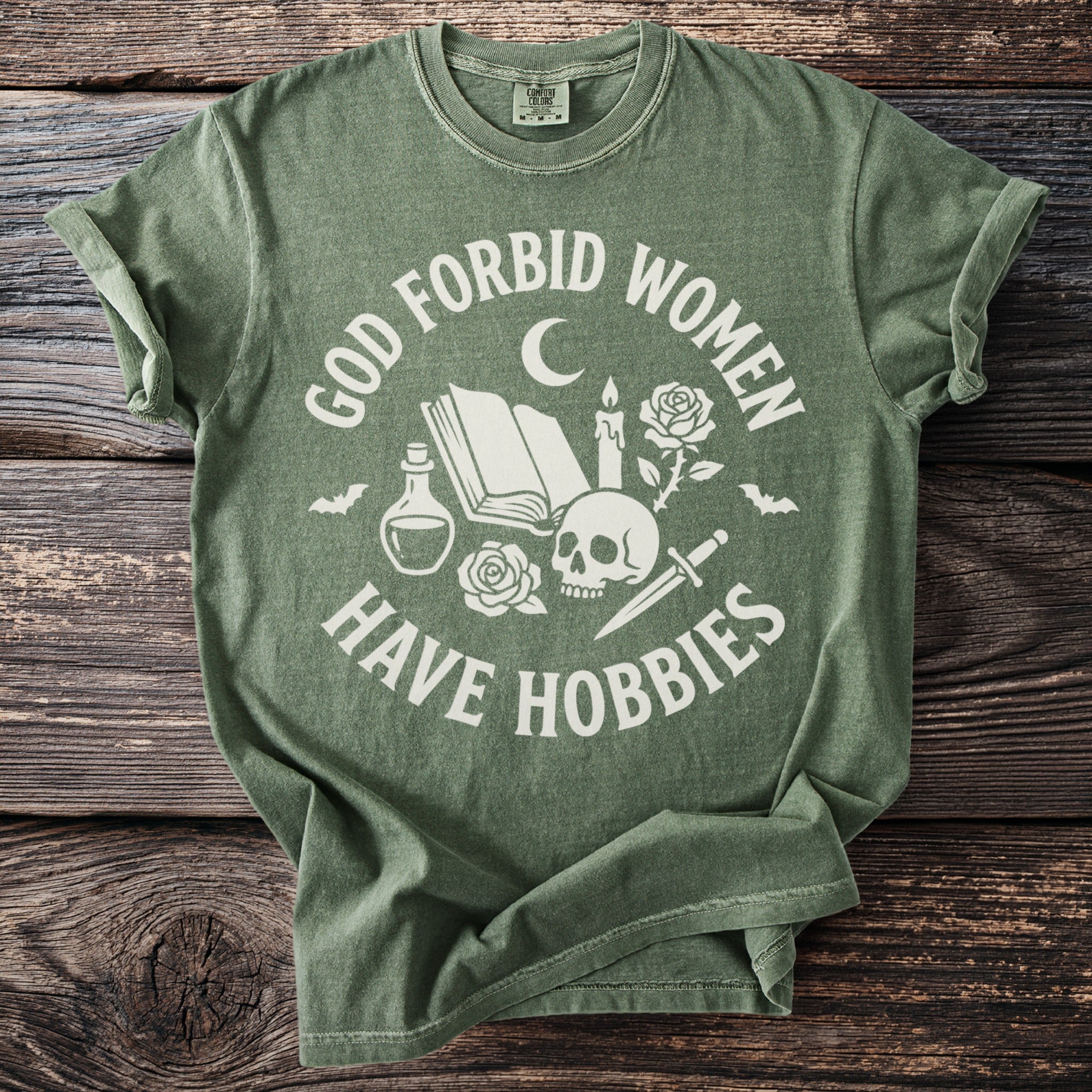 Green t-shirt with white text "God Forbid Women Have Hobbies" and gothic graphic elements.