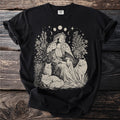 Illustration of a woman with cats, flowers, moon phases, and stars on a black t-shirt.