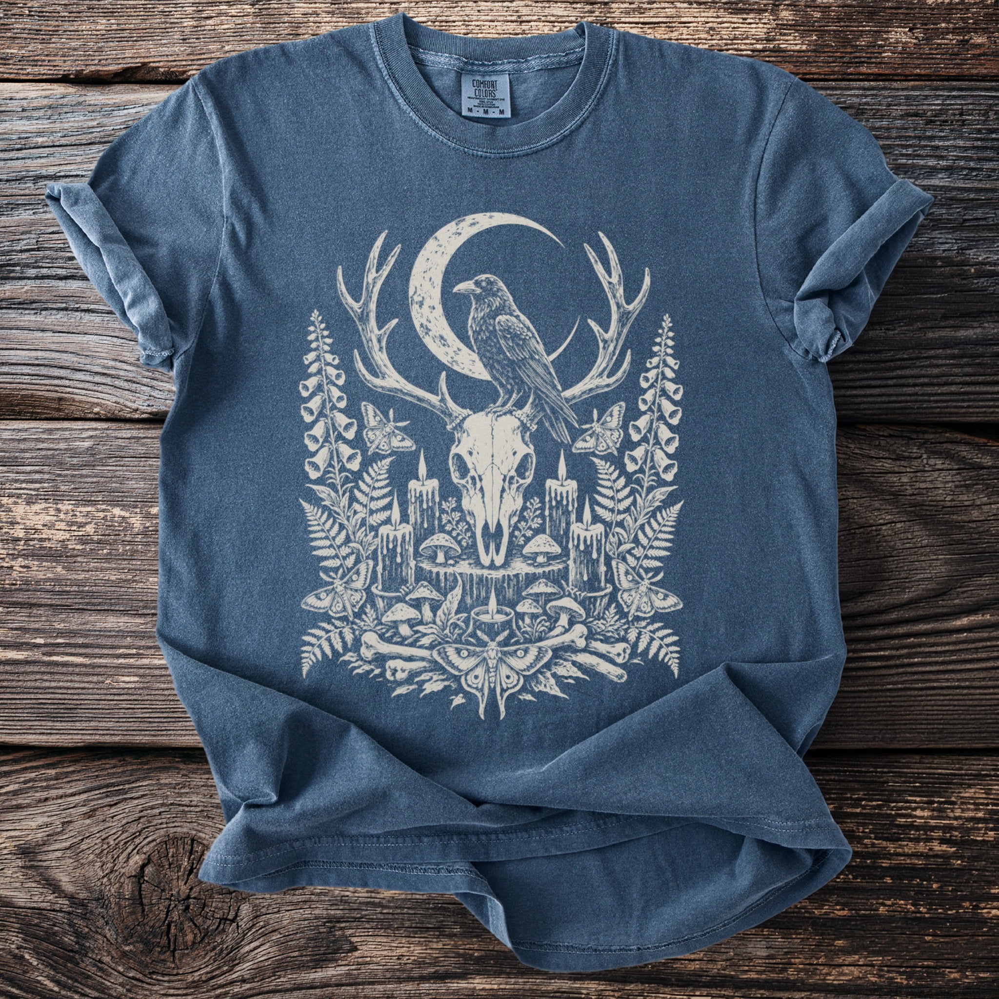 Blue t-shirt with cream graphic of a raven on deer antlers, skull, moon, and ornate nature elements.