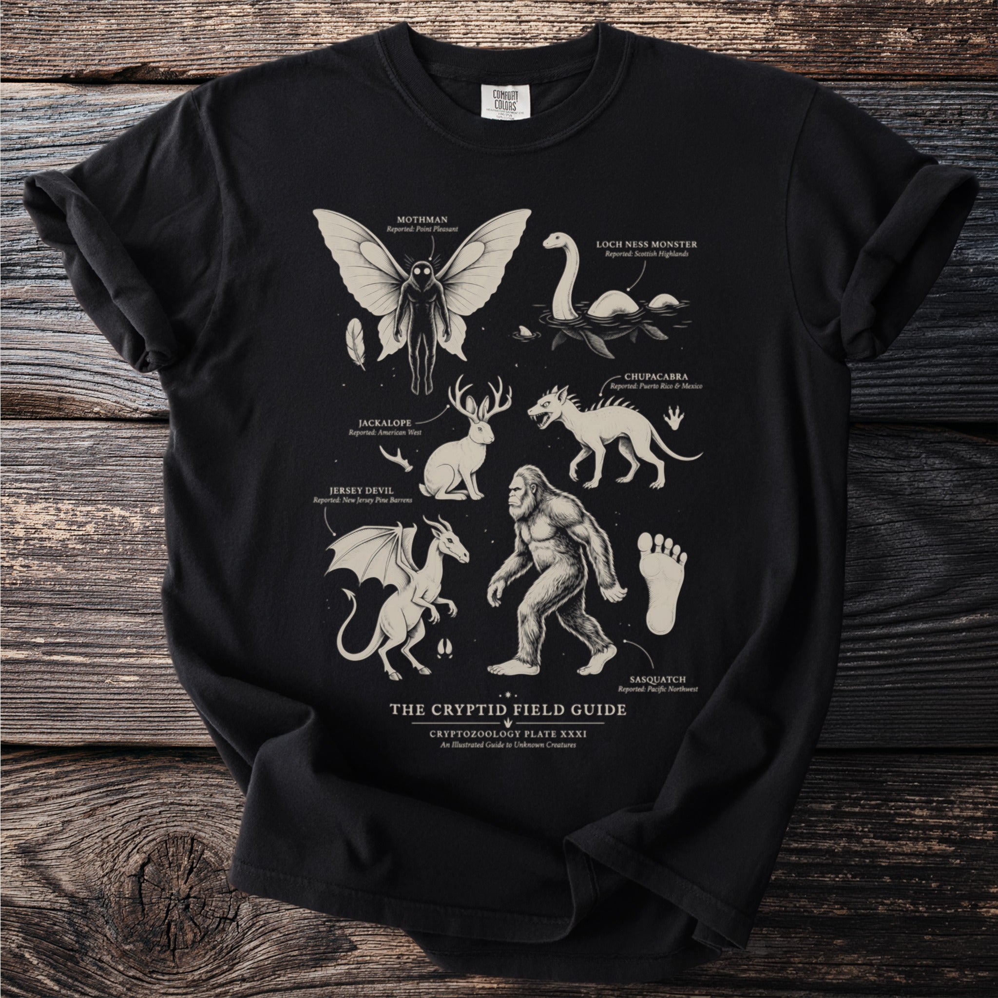 Black t-shirt featuring "The Cryptid Field Guide" with illustrations of Mothman, Nessie, Jackalope, Jersey Devil, Chupacabra, and Sasquatch.