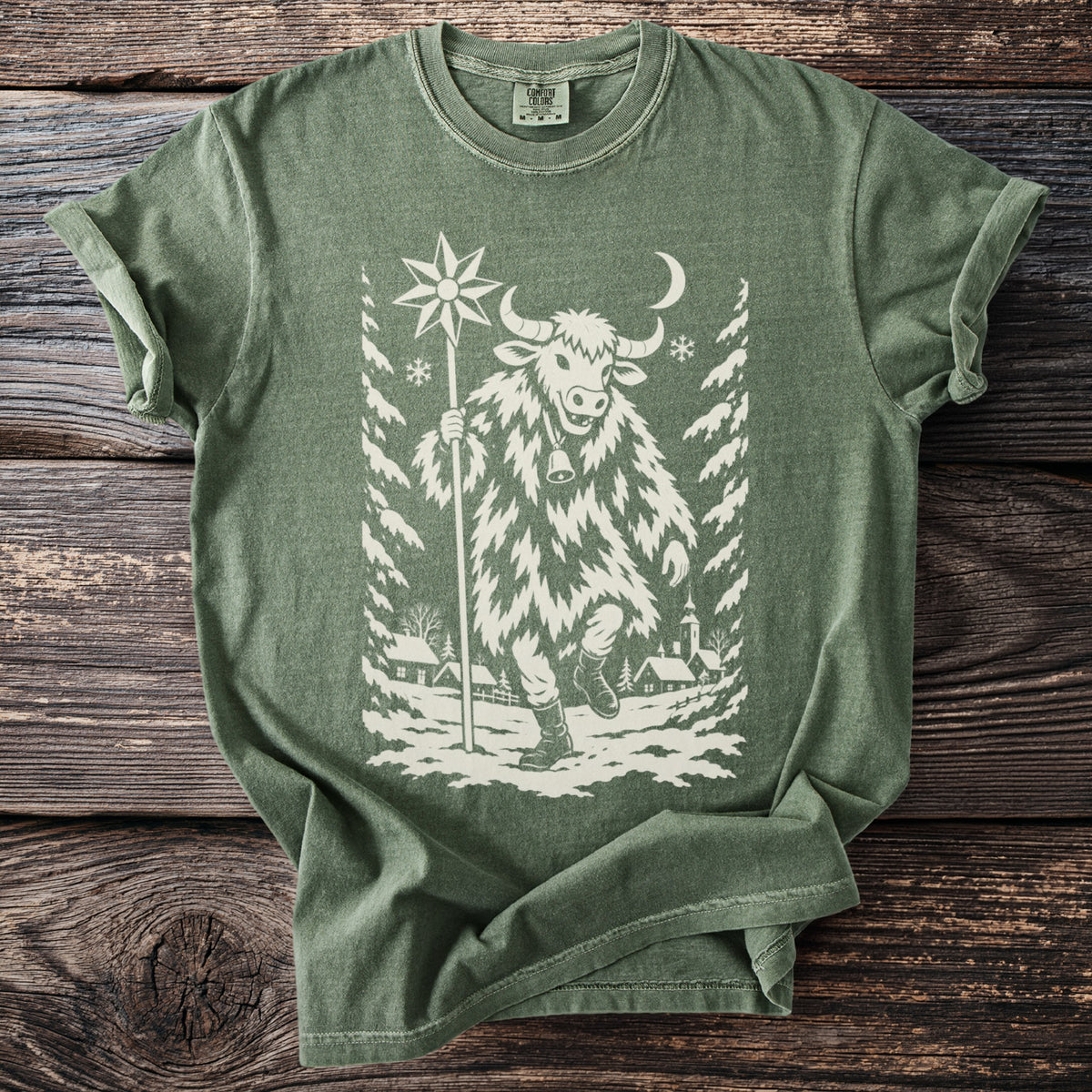 Moss green t-shirt with a cream folk-cut print: a bull-masked figure in a shaggy coat walks through a snowy village carrying a star on a pole.