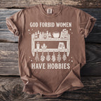 Brown t-shirt with "God Forbid Women Have Hobbies" and shelf graphic.
