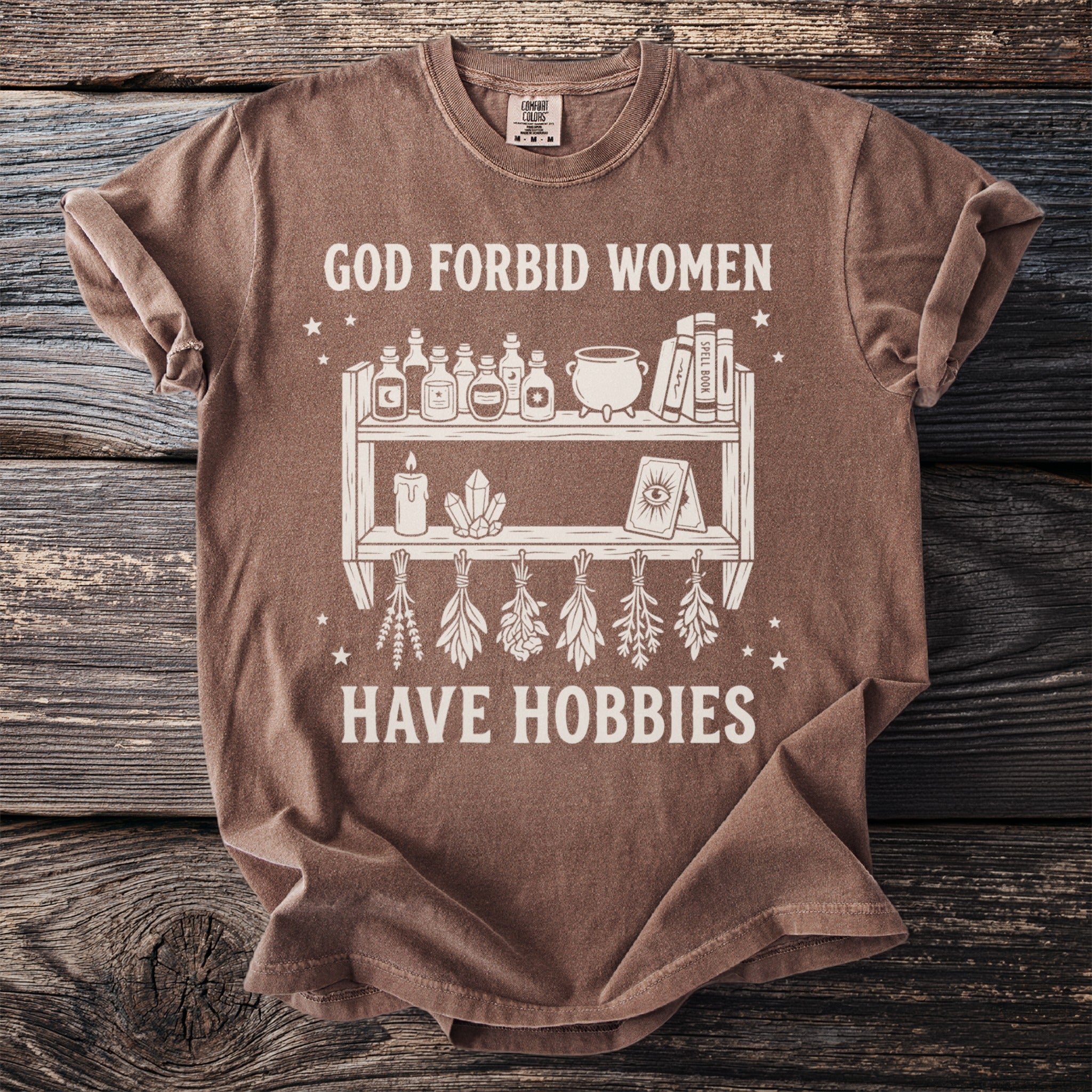 Brown t-shirt with "God Forbid Women Have Hobbies" and shelf graphic.
