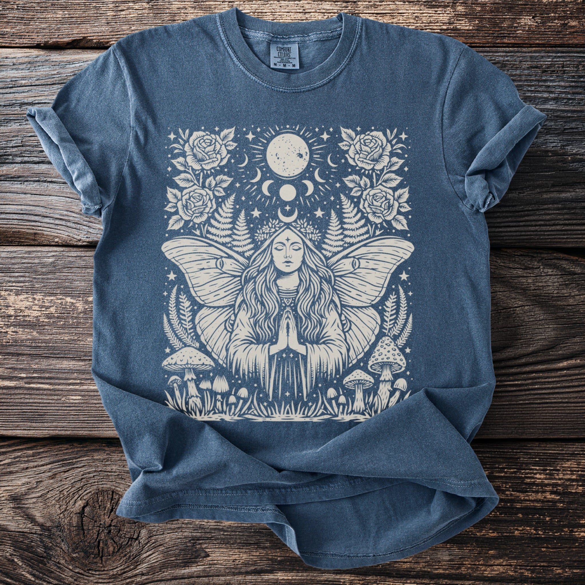 Blue t-shirt with a white moth woman, moon, stars, roses, ferns, and mushrooms design.