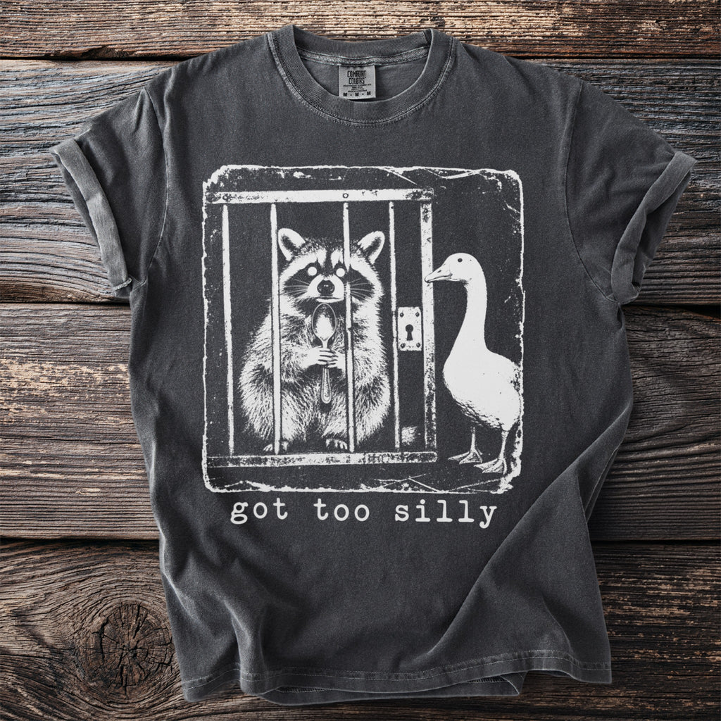 T-shirt with a graphic of a raccoon in a cage holding a spoon and a goose next to it with text "got too silly."