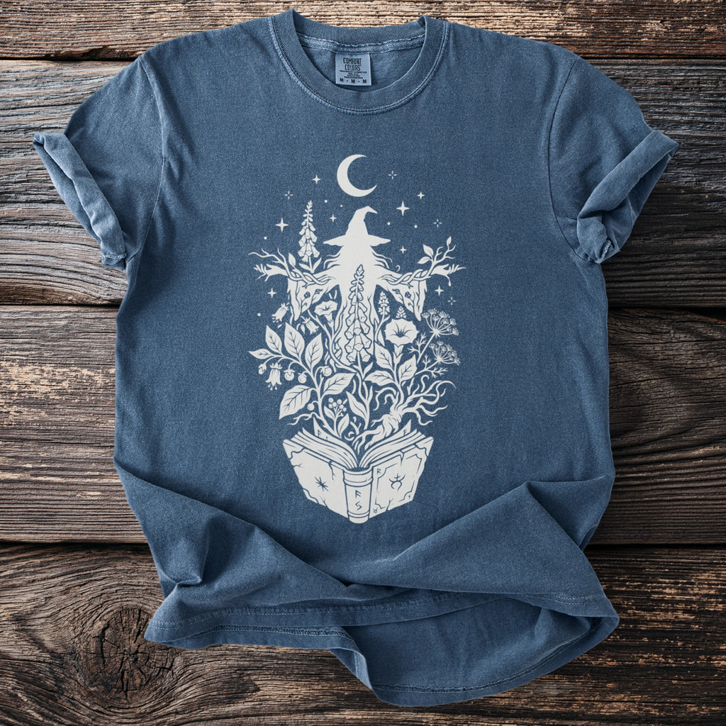 Blue t-shirt with white graphic of a witch surrounded by plants and a crescent moon.