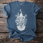 Blue t-shirt with white graphic of a witch surrounded by plants and a crescent moon.