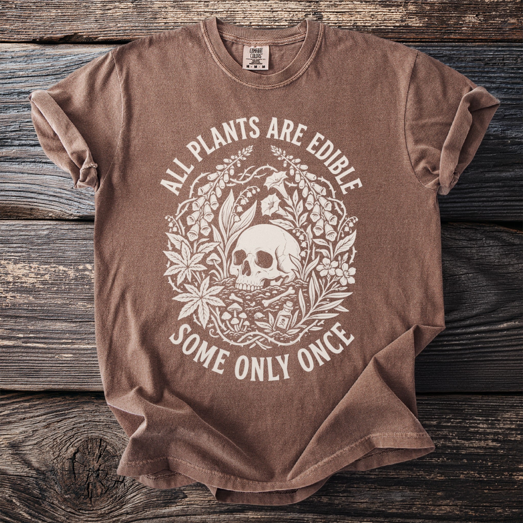 Brown t-shirt with a white circular graphic of a skull surrounded by plants and the text "ALL PLANTS ARE EDIBLE SOME ONLY ONCE".