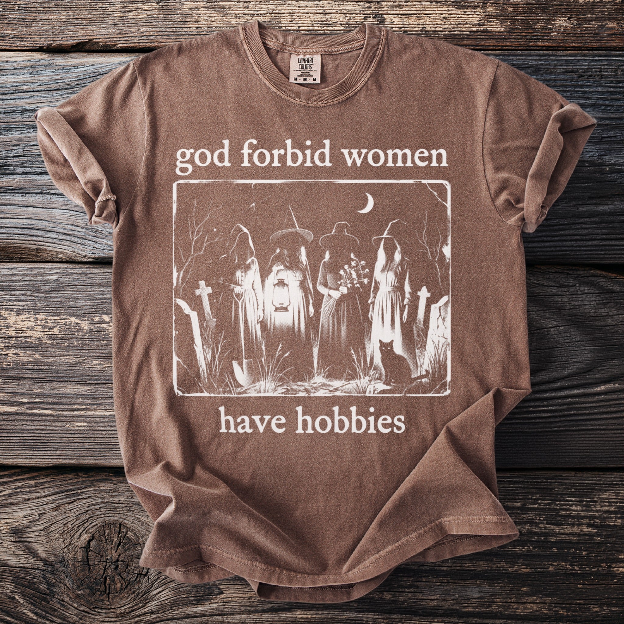 Brown t-shirt with "god forbid women have hobbies" text and witch illustration.