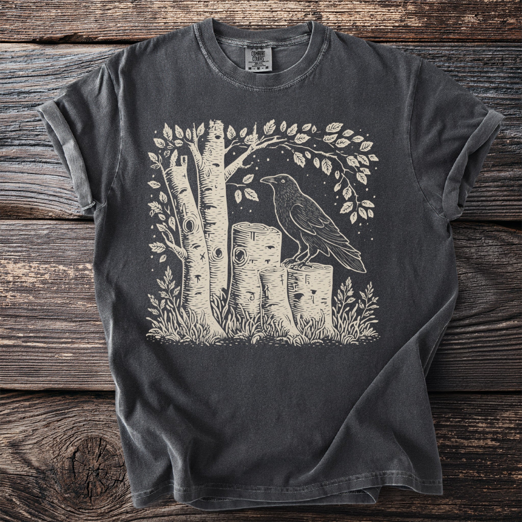 T-shirt with a linocut-style print of a raven on logs among birch trees.
