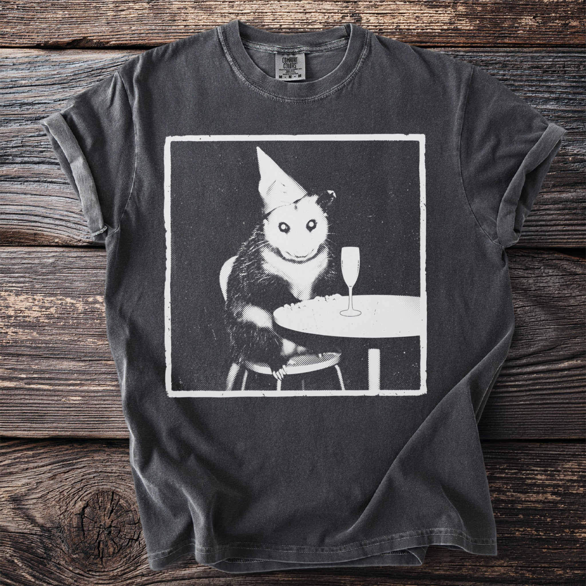 Black t-shirt with a vintage print of a possum wearing a party hat at a table.