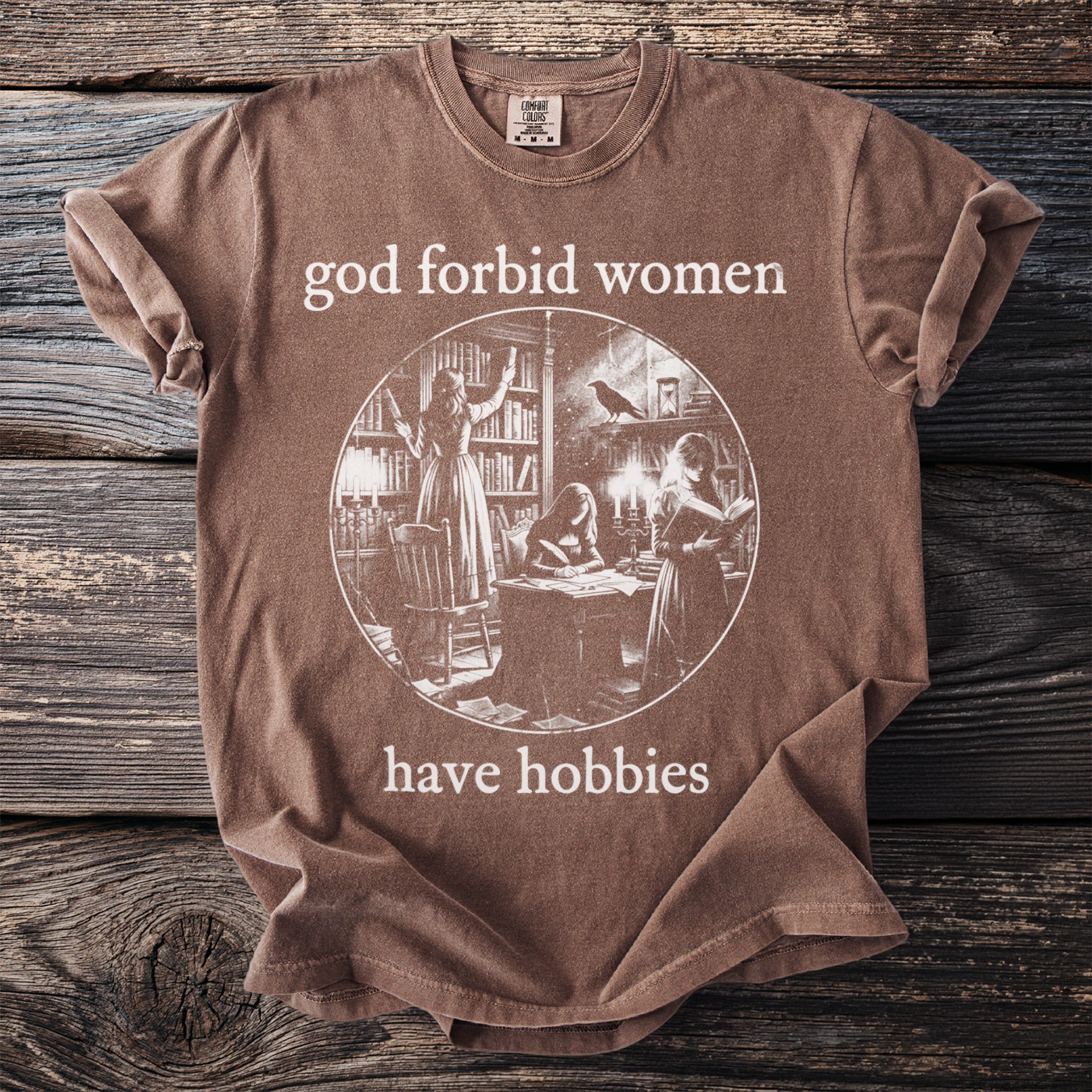 Brown t-shirt with text "god forbid women have hobbies" and library illustration.