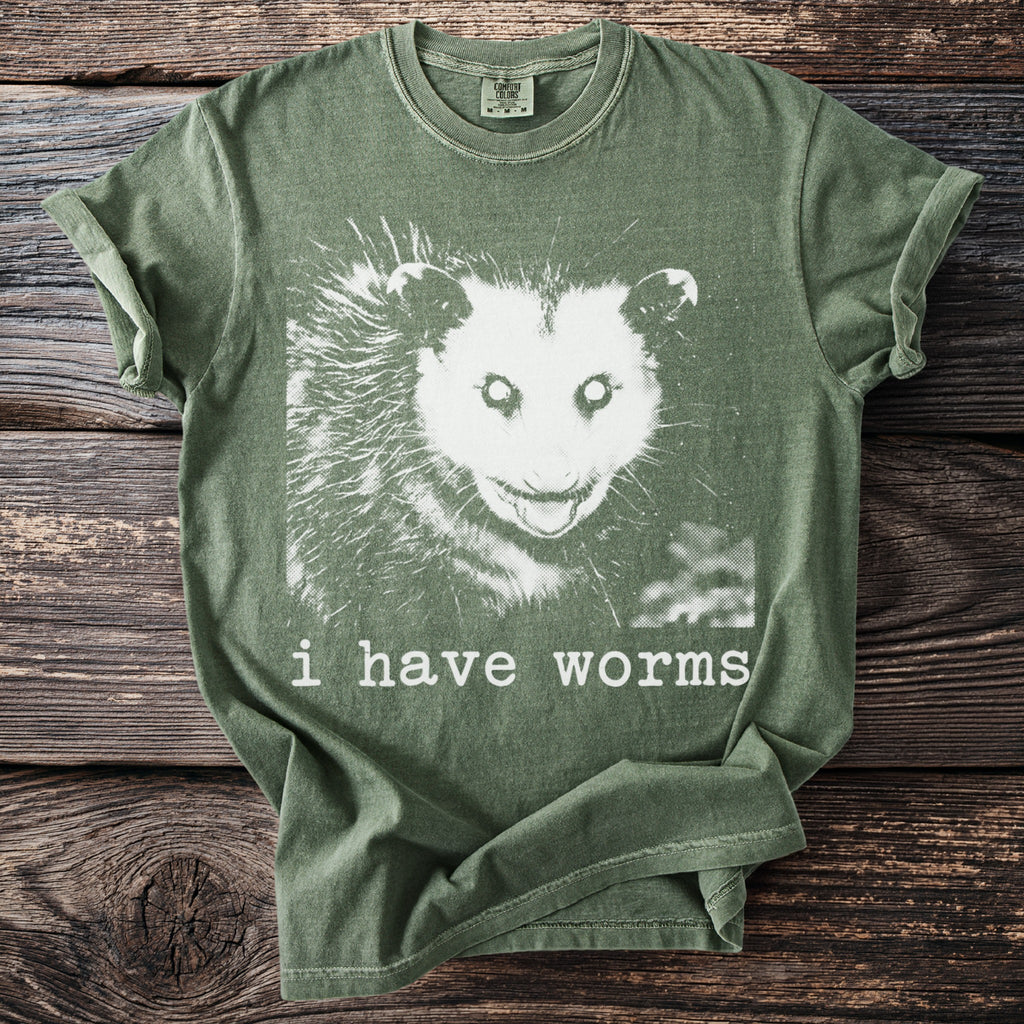 Green t-shirt with a white graphic of an opossum face and text "i have worms".