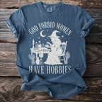 T-shirt with white graphic of witch at a table and text "GOD FORBID WOMEN HAVE HOBBIES".