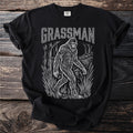 Black t-shirt with white graphic of Bigfoot walking through reeds and water, text reads "GRASSMAN".