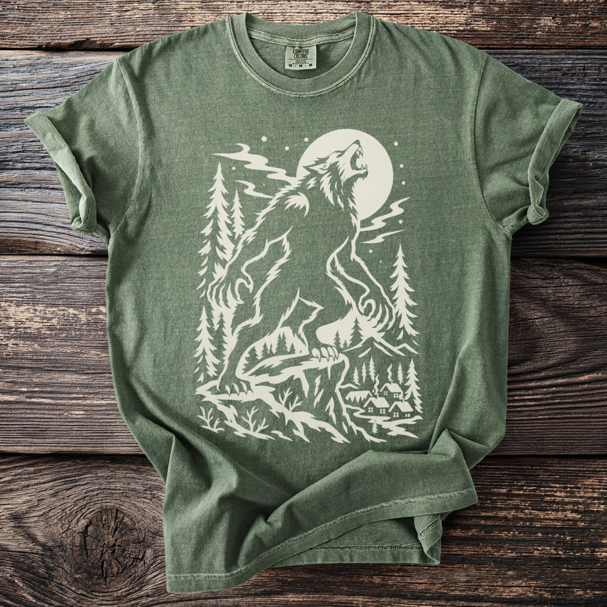 Green t-shirt with cream graphic of a wolf howling at the moon in a forest with village.