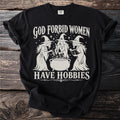 Black t-shirt with white text "God Forbid Women Have Hobbies" and three witch illustrations.