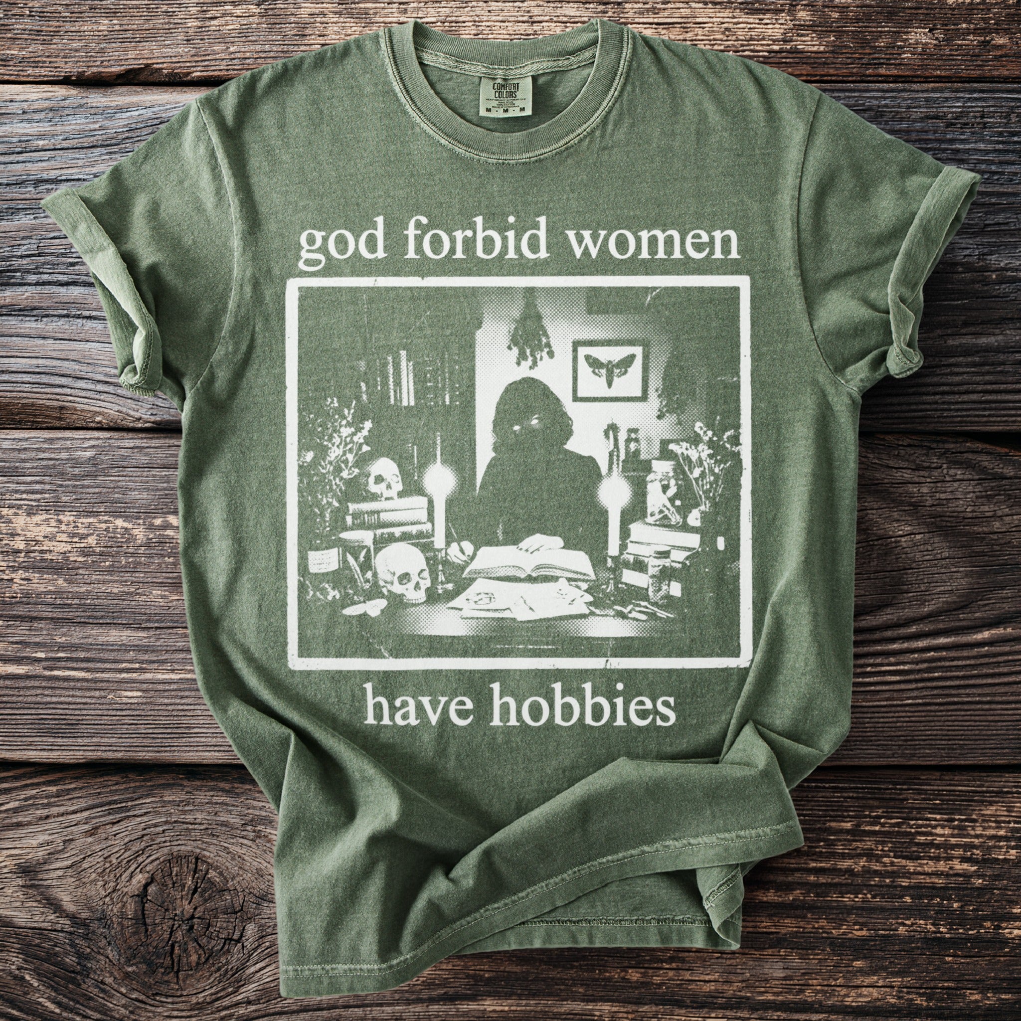 Green t-shirt with "god forbid women have hobbies" text and gothic illustration.