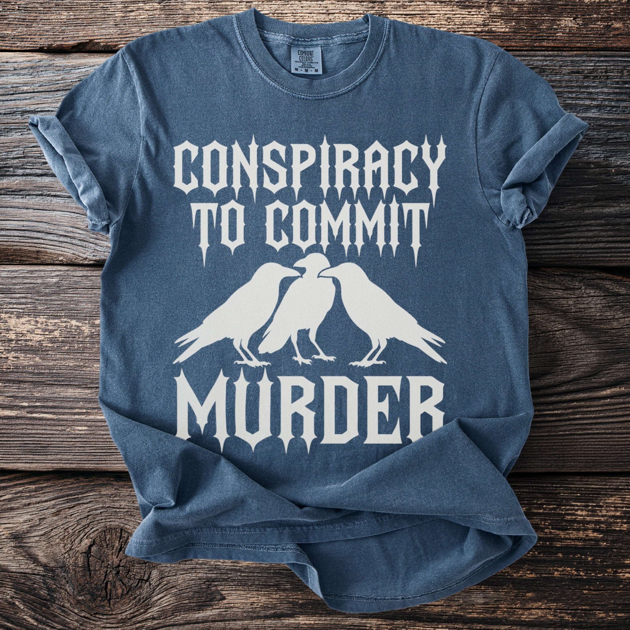 Blue t-shirt with white text "Conspiracy to commit murder" and three crows.