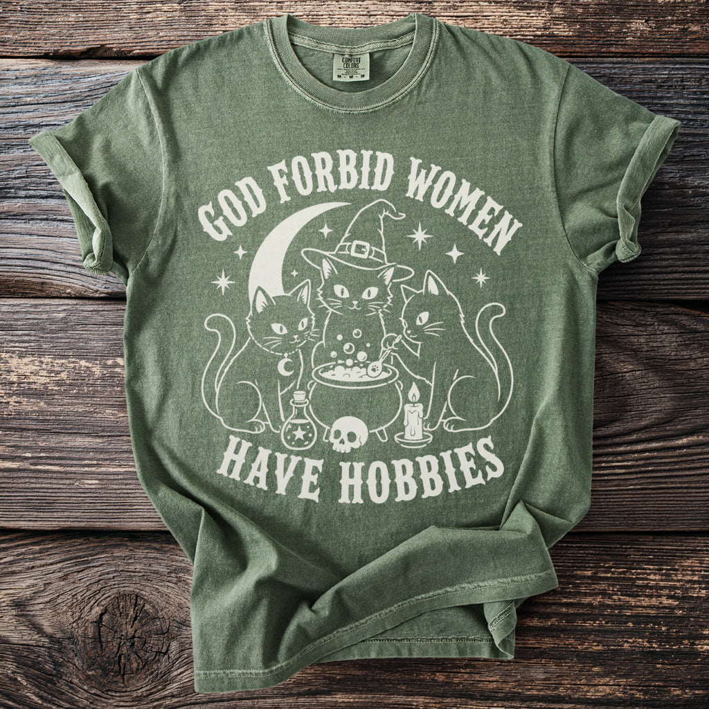 Green t-shirt with white graphic of cats, cauldron, skull, and text "God Forbid Women Have Hobbies".