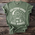Green t-shirt with white graphic of cats, cauldron, skull, and text "God Forbid Women Have Hobbies".