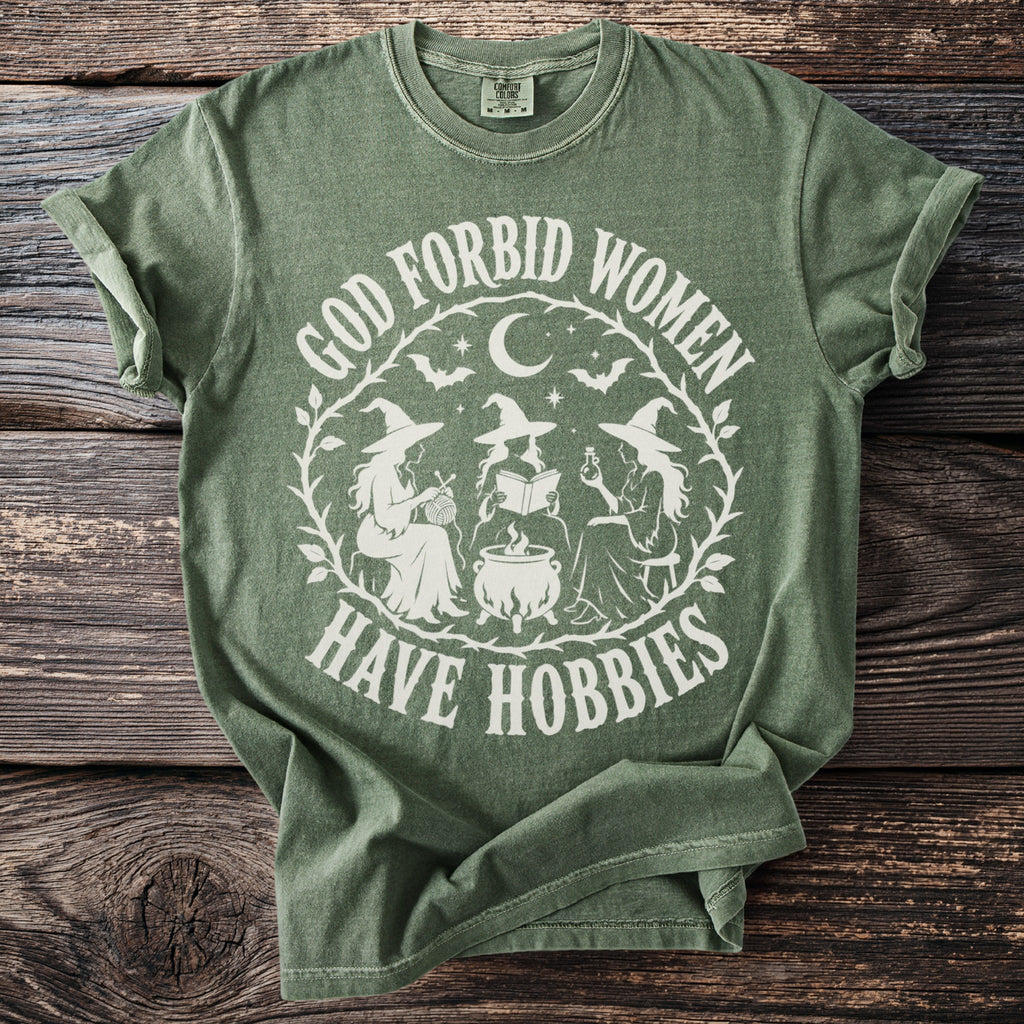 Green t-shirt with white graphic of three witches, cauldron, moon, bats, and text "God Forbid Women Have Hobbies."