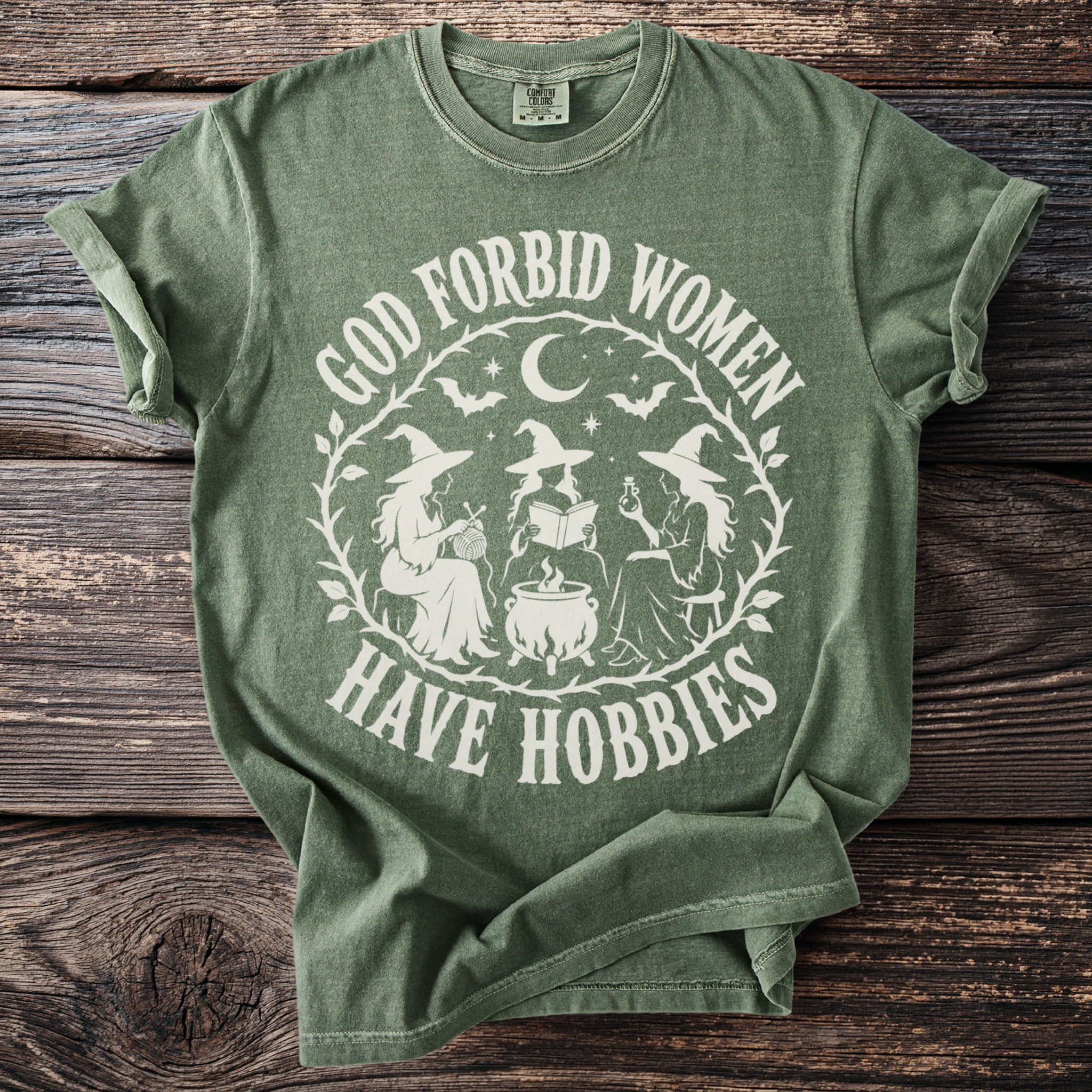 Green t-shirt with white graphic of three witches, cauldron, moon, bats, and text "God Forbid Women Have Hobbies."