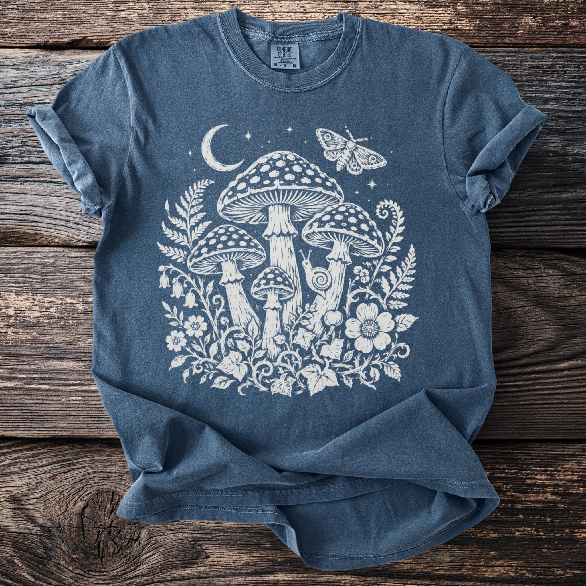 Blue t-shirt with white graphic of mushrooms, moon, stars, butterfly, and snail.