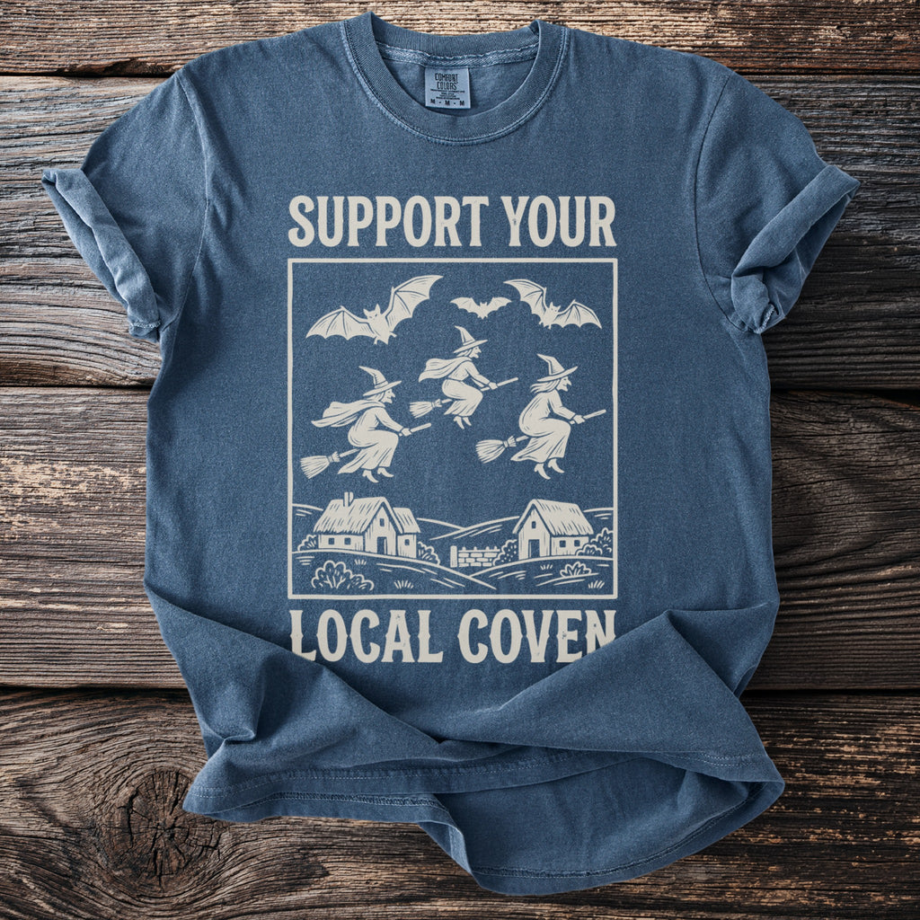 T-shirt with white graphic of witches flying on brooms over houses, and bats, reading "Support Your Local Coven".